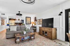 A living room with a sofa, coffee table, TV, wooden cabinet, and artwork on the wall. - Aldea 1, Belleair Beach, 2BR,2BA (Belleair Beach)