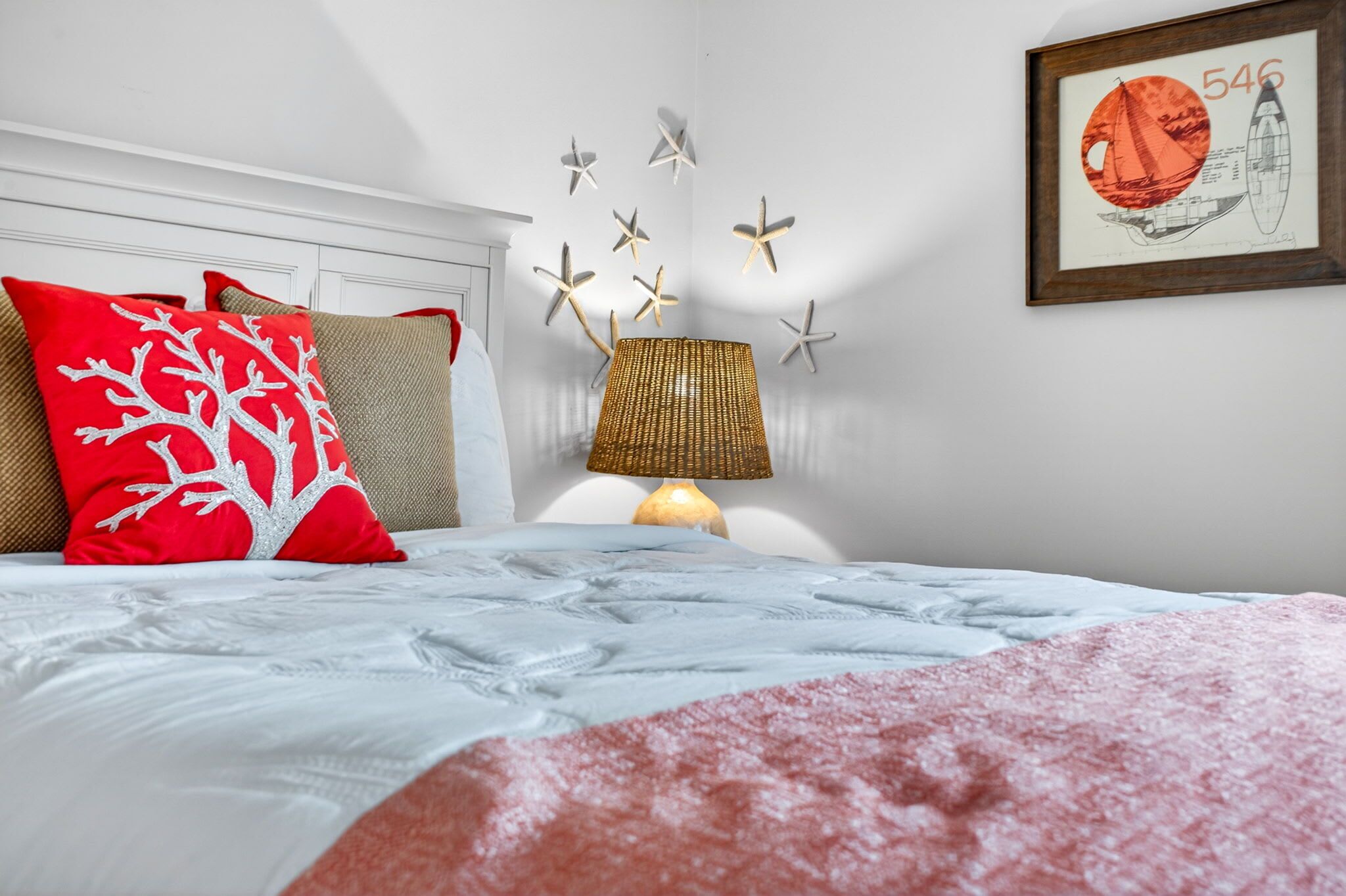 Ein ordentlich bezogenes Bett mit einer weißen und roten Decke, einer Korbtasche als Lampe sowie Wanddekoration mit Meeressternnen.
