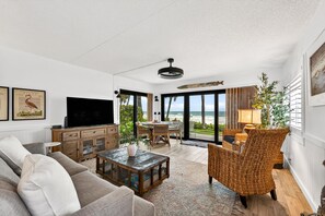 A living room with a sofa, coffee table, TV, wooden cabinet, wicker chair, and a view of the beach through large windows. - Aldea 1 Beachfront Condo in Belleair Beach (Belleair Beach)