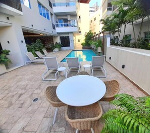 Uma área de piscina ao ar livre com espreguiçadeiras e uma mesa, rodeada por edifícios e vegetação.