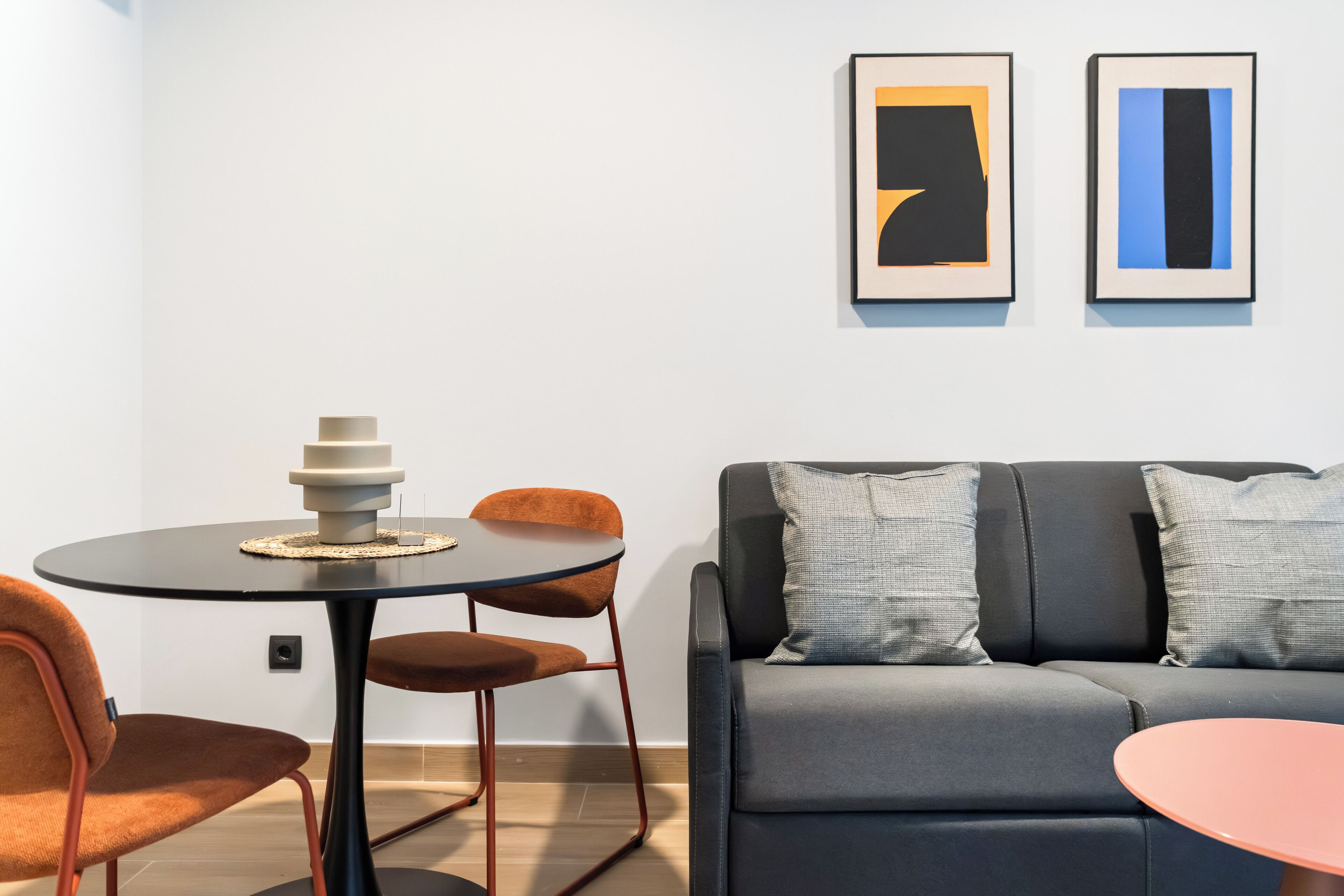 A modern living room with a grey sofa, orange chairs, a small round table with a stack of books, and two abstract art pieces on the wall.