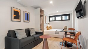 Un salon moderne avec un canapé noir, une table ronde et des chaises orange. On y trouve un lit avec une literie blanche et une fenêtre avec des stores.