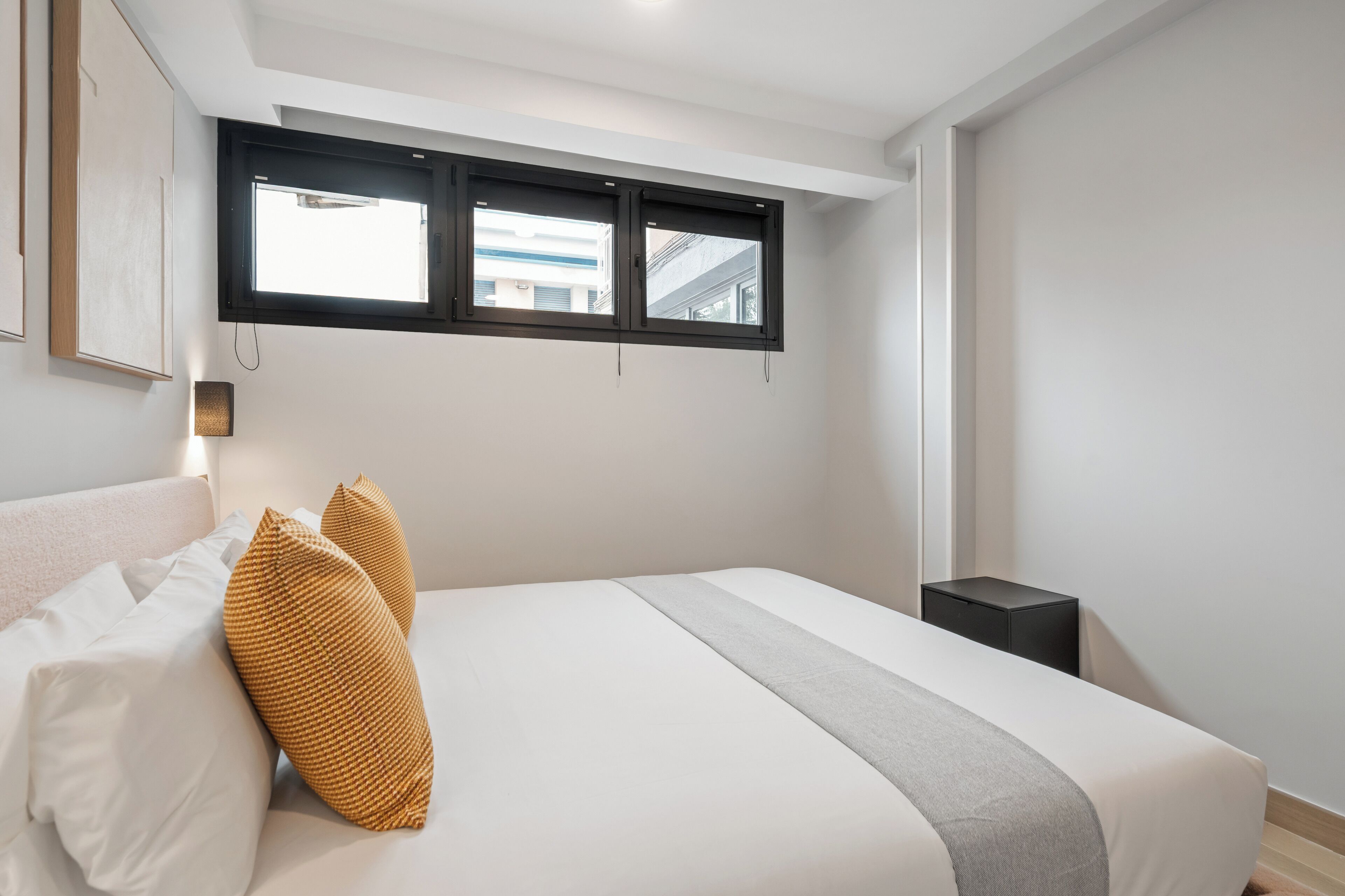 A modern bedroom with a large bed, two windows, a bedside table, and a wall-mounted light.