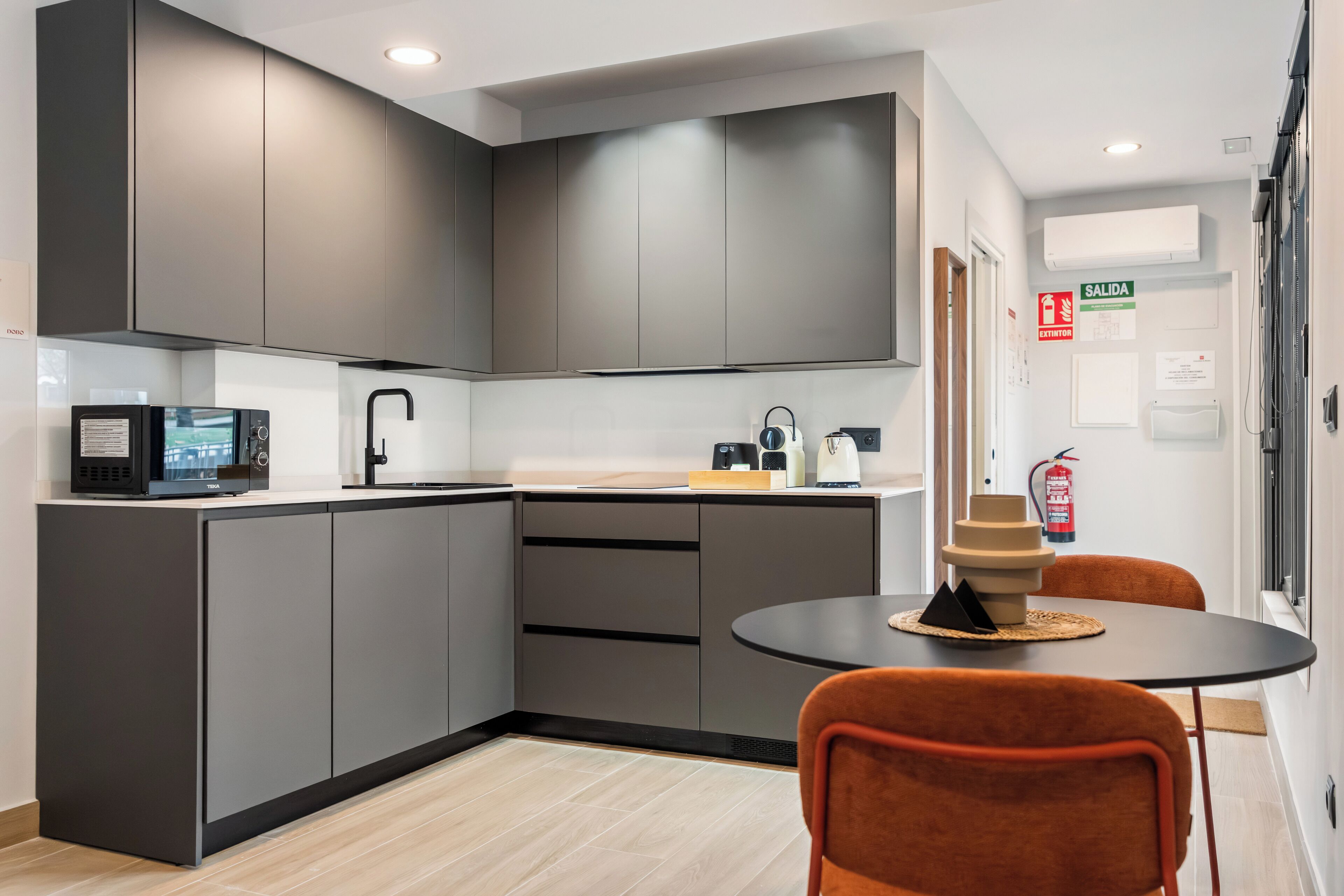 A modern kitchen with grey cabinets, a black countertop, and a round table with two orange chairs.