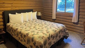 Un dormitorio tipo cabaña de madera con una cama, una mesita de noche, una lámpara y una ventana con cortinas.