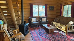 A log cabin interior with a fireplace, two armchairs, a sofa, a coffee table, and a patterned rug.