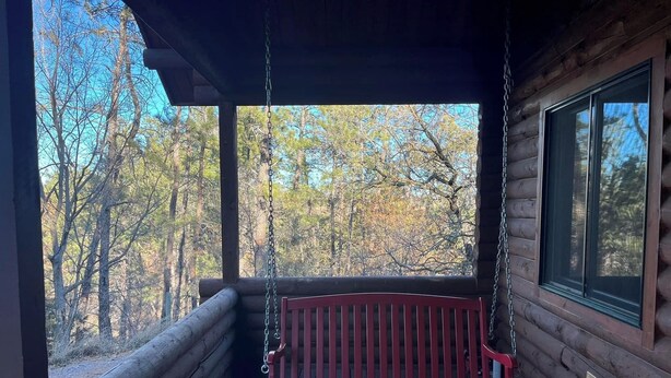 A log cabin with a wooden porch, a red swing, and a view of a forest.