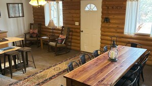 Interior de una cabaña de madera con una mesa de comedor grande, sillas, una alfombra y una puerta.