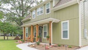 A two-story house with a covered porch, a number '210' on the door, and a landscaped front yard with trees and shrubs. - Cozy New House in a Prime Location (Carrollton)