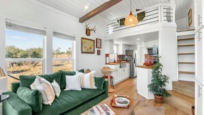 A spacious living area with a green sofa, a coffee table with books, and a view of the outdoors. - Secluded Hill Country Ranch on 105 Acres Near Pedernales State Park (Johnson City)