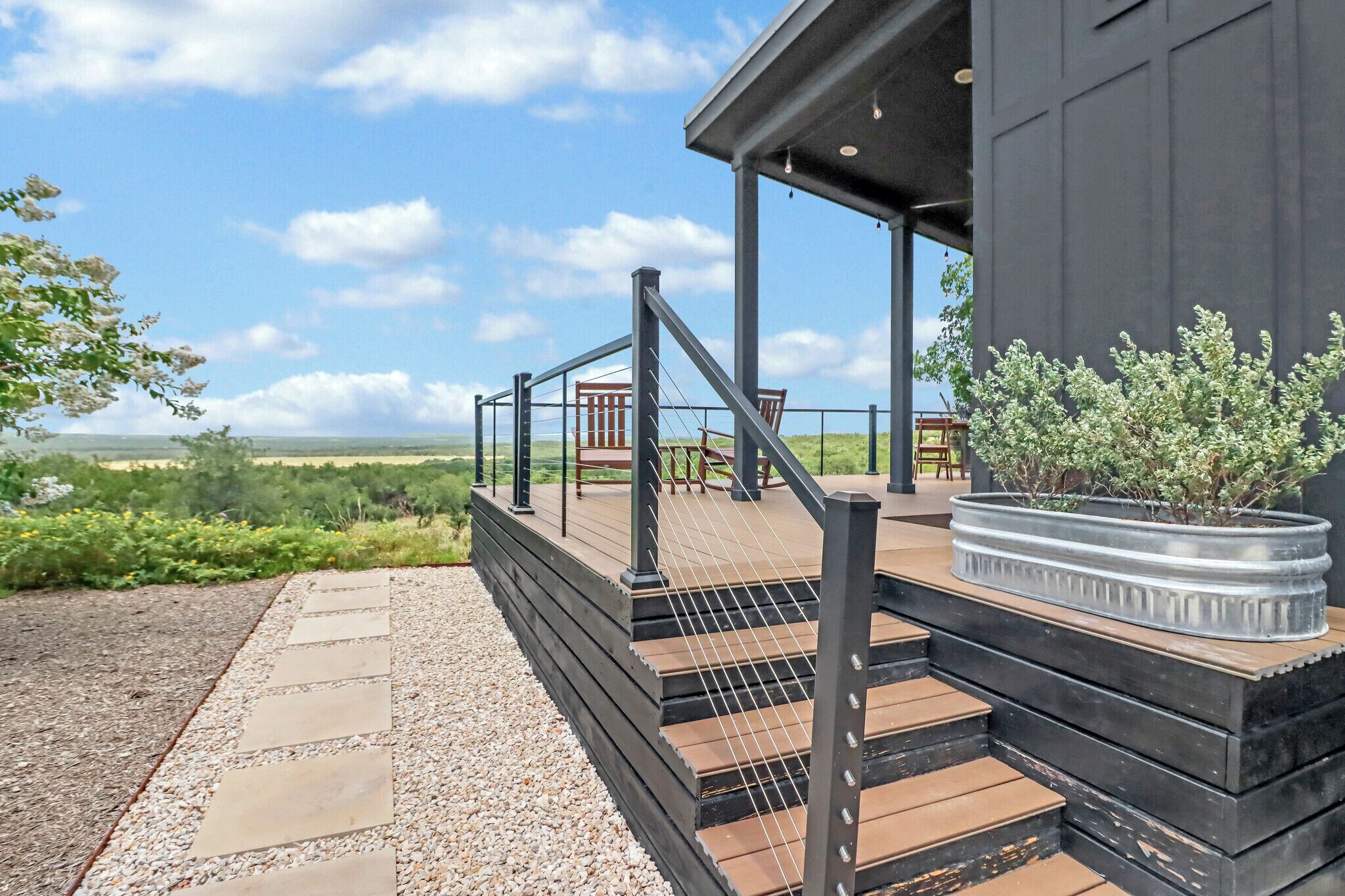 A modern outdoor deck with steps leading to a covered area, surrounded by a gravel path and greenery.
