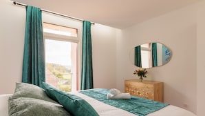 A bedroom with a bed, teal curtains, a wooden dresser, a mirror, and a window with a view of greenery.