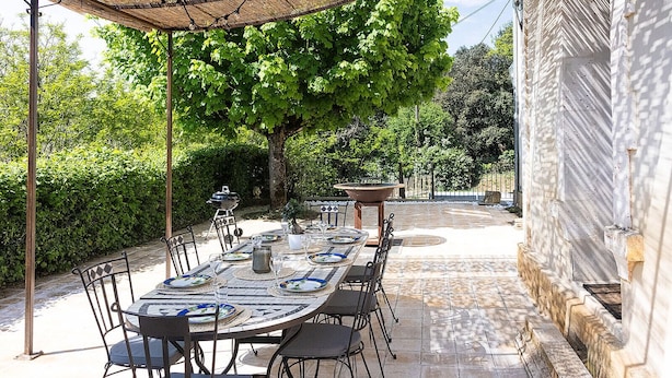 An outdoor dining area with a long table set for six, surrounded by chairs, under a shaded pergola.