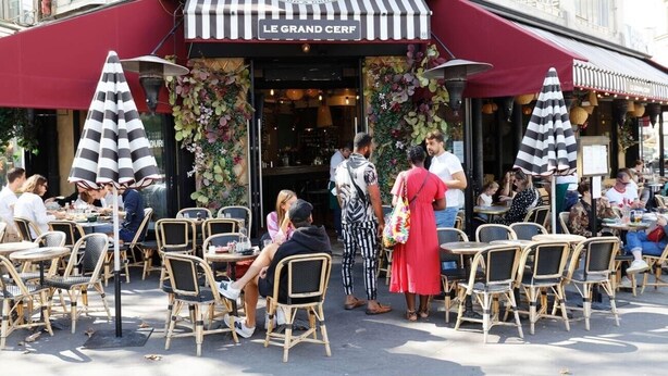 Un caffè all'aperto con sedie e tavoli in vimini, una tenda a righe e un cartello con scritto “LE GRAND CERF”.