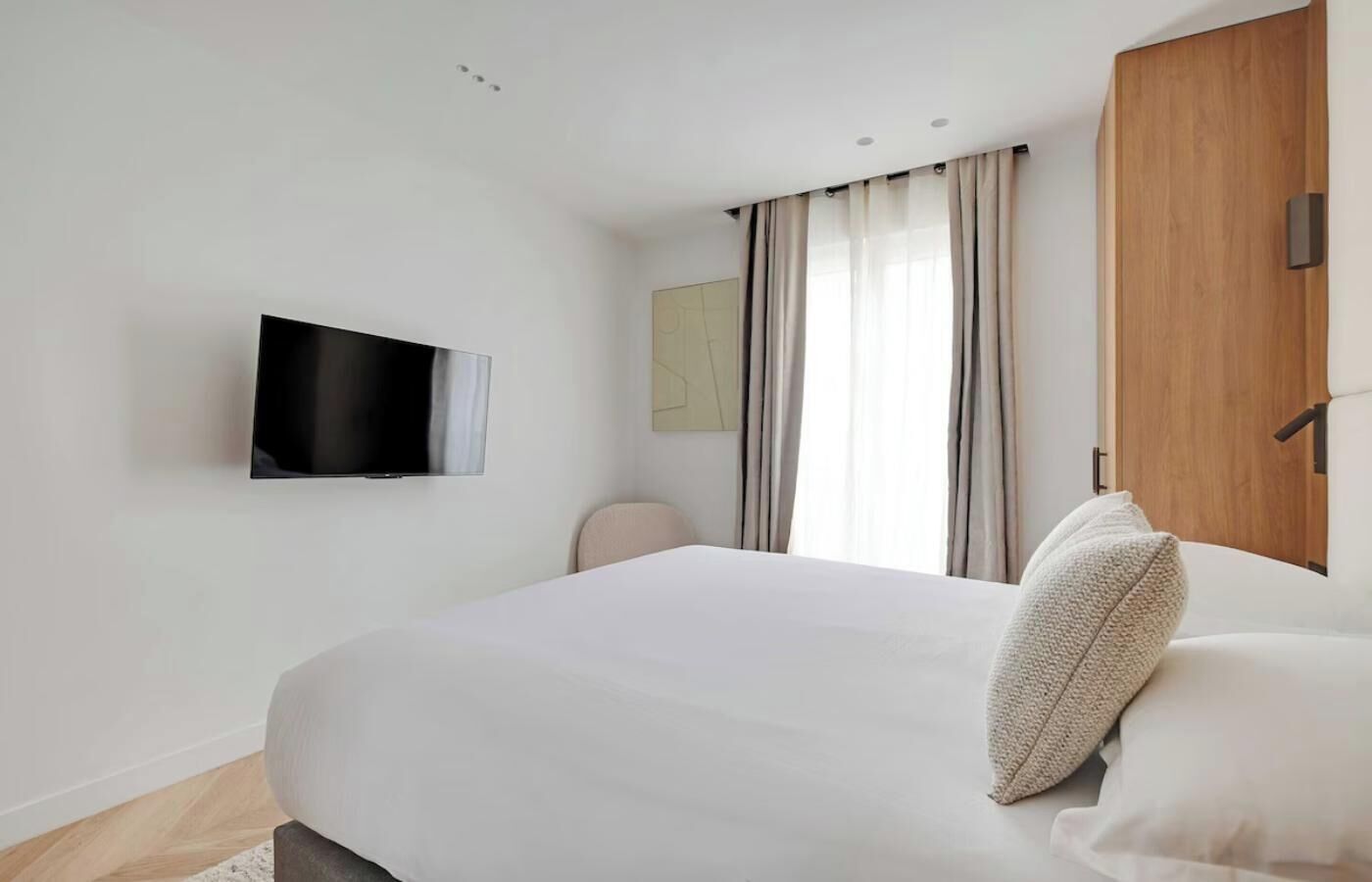 Ein modernes Hotelzimmer mit einem großen Bett, einem flachbildfernseher an der Wand, einem Holzschrank und einem Fenster mit Vorhängen.