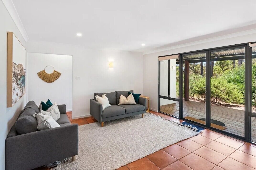A modern living room with a grey sofa, a smaller grey armchair, and a white rug. A large window offers a view of greenery outside.