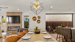 A modern dining area with a long table set for six, a TV mounted on the wall, and a fireplace. - Simply Paradise: Steps to Desert Color Lagoon + Beaches (St. George)