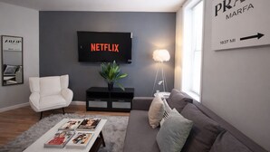 Una sala de estar moderna con un sofá gris, un sillón blanco, una mesa de centro con revistas, un televisor mostrando 'NETFLIX' y un cartel con la inscripción 'PRADA MARFA' en la pared.
