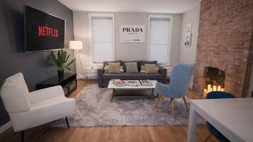 A modern living room with a flat-screen TV, a grey sofa, a white armchair, a coffee table with magazines, and a brick fireplace.
