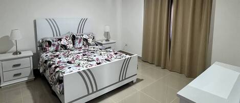 A bedroom with a bed, bedside tables, an air conditioning unit, a window with curtains, and a small white cabinet.
