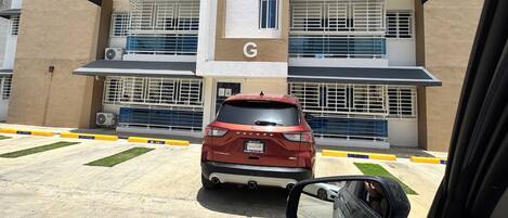 Ein mehrstöckiges Gebäude mit einem markanten 'G' auf der Fassade, Klimaanlagen und einem roten Auto vor der Tür.