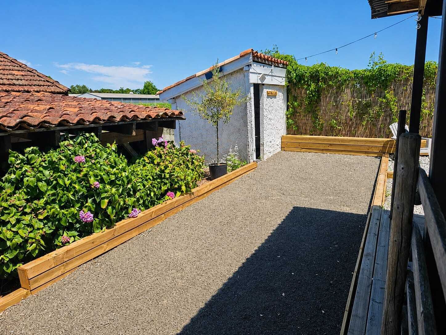 A gravel pathway leading to a building with a tiled roof, surrounded by wooden raised beds with green plants.