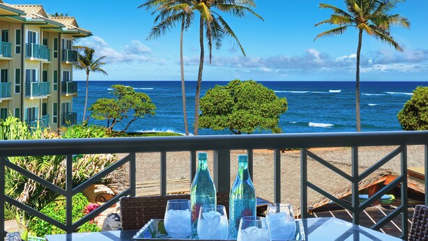A balcony with a table set for drinks, overlooking a beach with palm trees and ocean waves. - Waipouli Beach Resort Exquisite Ocean & Pool View - Kauai Luxury with 2 Kings (Kapaa)