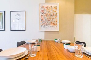 Un coin repas avec une table en bois dressée pour quatre personnes, des œuvres d’art encadrées accrochées au mur et une porte blanche en arrière-plan.