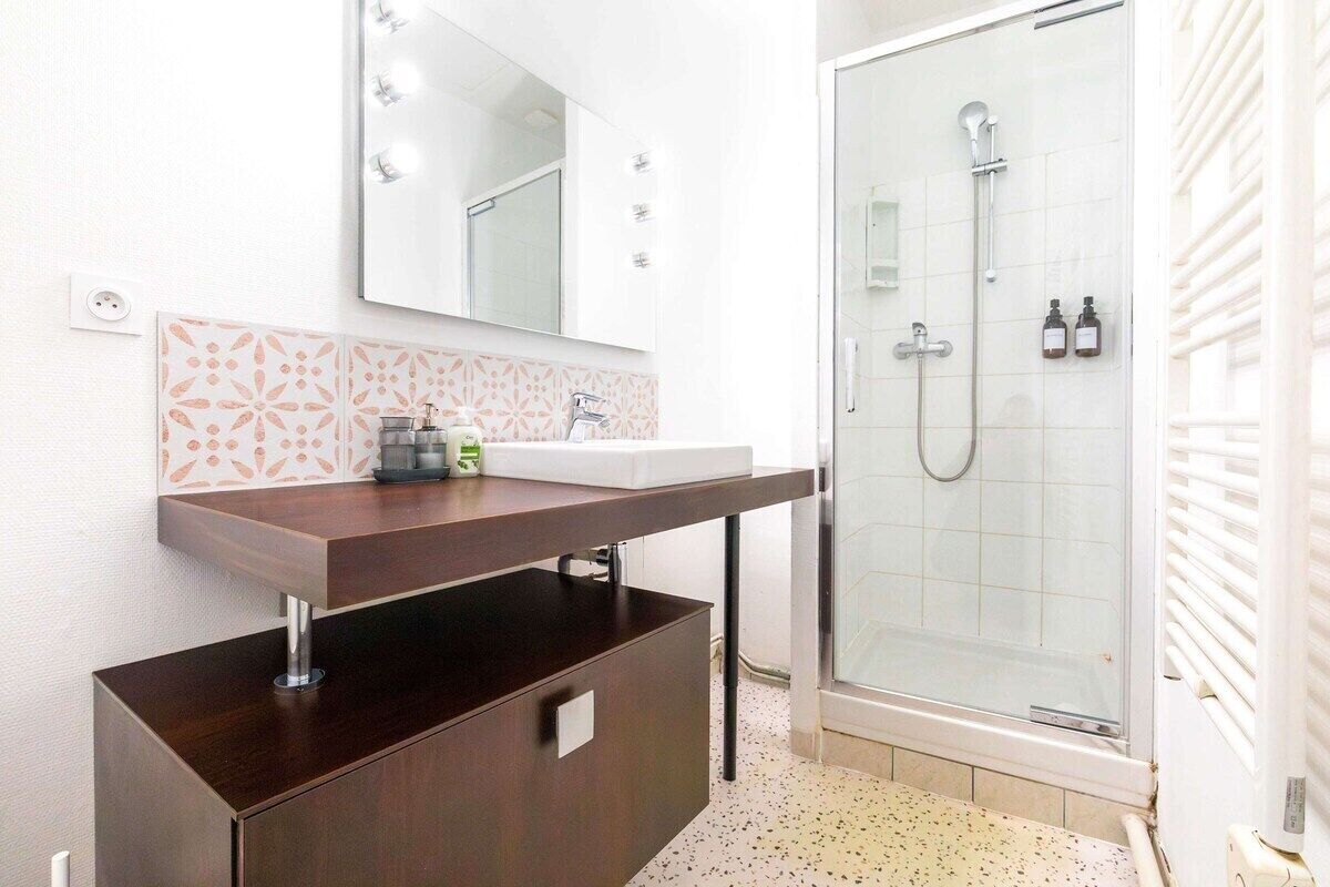 Un baño con tocador de madera oscura, lavamanos blanco, espejo grande y cabina de ducha de vidrio.