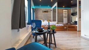 A modern hotel room with a dining area, a bed with a white headboard, a small table with a plant, and a blue chair. - 🛁 Balnéo Suite Arquebuse — Love'appart & Jacuzzi (Dijon)