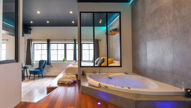 A modern bathroom with a large bathtub, a mirror, and a wooden floor. - 🛁 Balnéo Suite Arquebuse — Love'appart & Jacuzzi (Dijon)