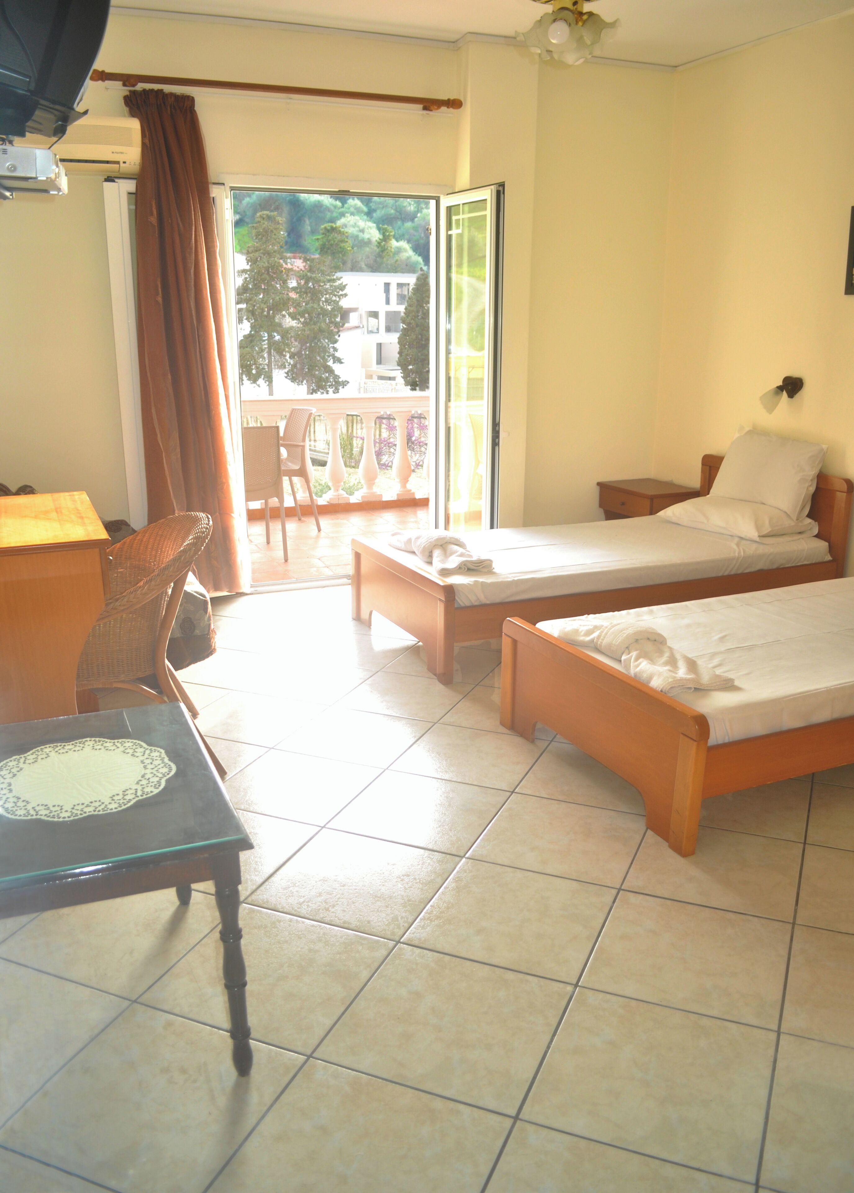 Ein Hotelzimmer mit zwei Betten, einem Schreibtisch, einem Sessel und einem Balkon mit Blick auf Bäume und Gebäude.