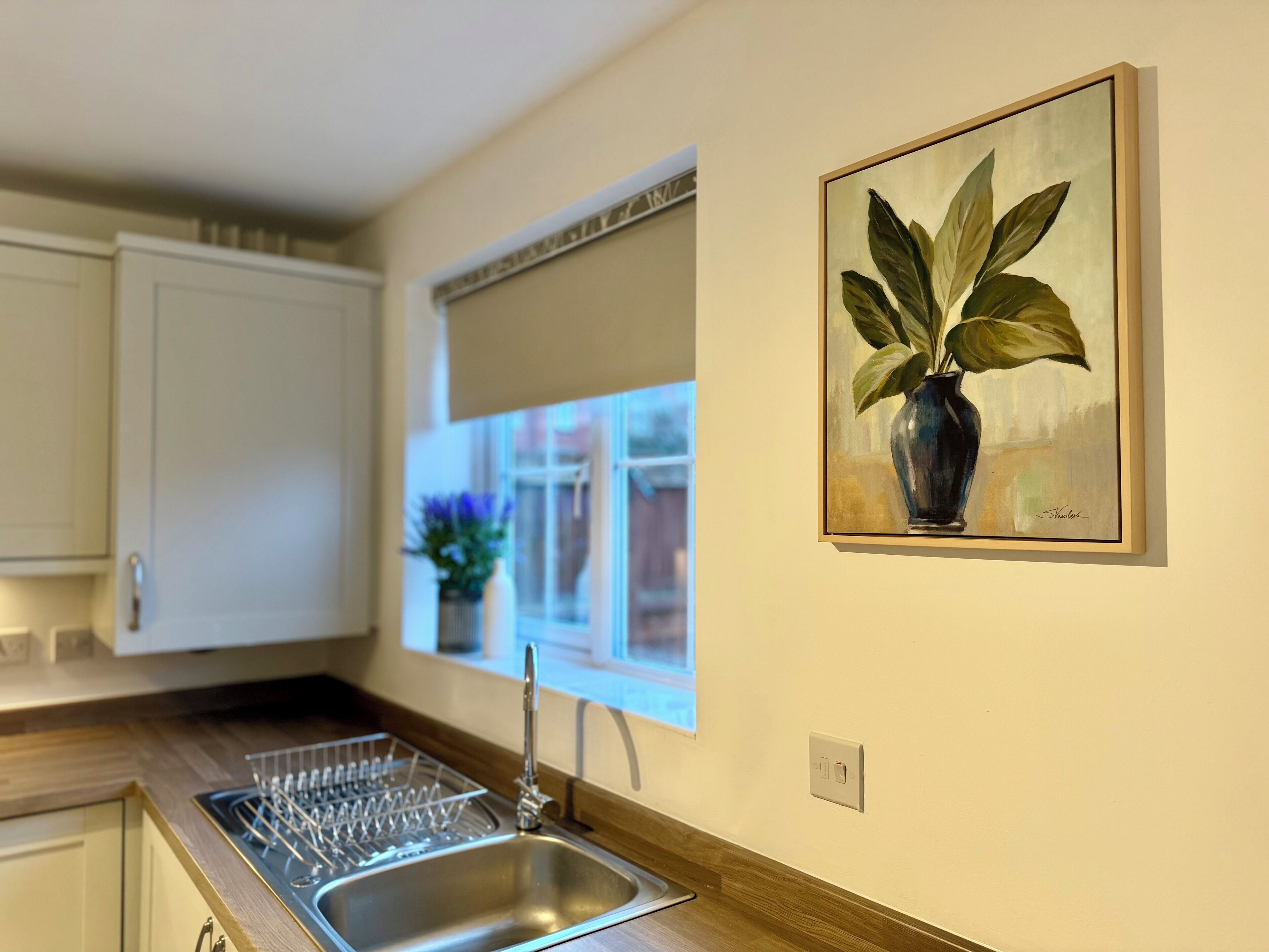 A kitchen with a stainless steel sink, a window with a view, and a painting of a plant on the wall.