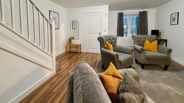 A living room with a staircase, grey armchairs with yellow cushions, a wooden floor, and a ceiling light fixture. - 4 Bed | Free Parking | Fast WiFi | Desk | Pets (Buckshaw Village)