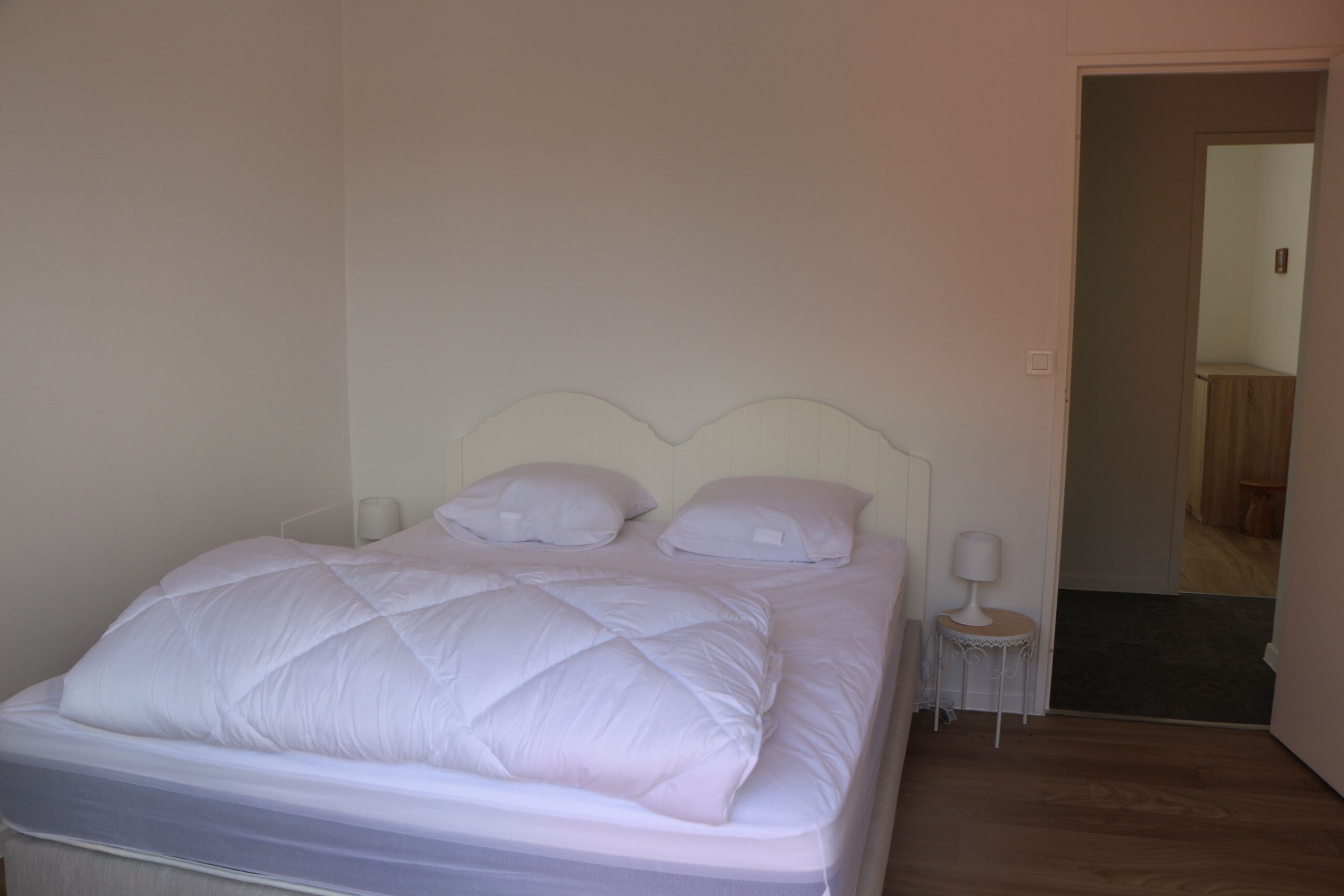 A neatly made bed with white bedding and pillows, a bedside table with a lamp, and a door leading to another room.