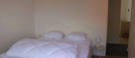 A neatly made bed with white bedding and pillows, a bedside table with a lamp, and a door leading to another room.
