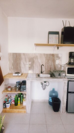 A compact kitchen with a sink, a microwave, and a water dispenser. There are shelves with various items and a trash bin.
