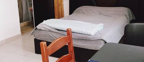 A room with a bed, a table, a chair, an air conditioning unit, and a door leading to a bathroom.