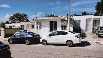 Un carrer residencial amb dos cotxes aparcats davant de cases, cel blau i una antena parabòlica visible en un dels edificis.