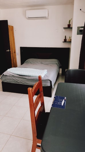 A room with a bed, a table, a chair, an air conditioner, and a door.