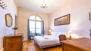 Habitación de hotel con cama, escritorio, silla, cómoda, candelabro, ventana con cortinas y un cuadro en la pared.