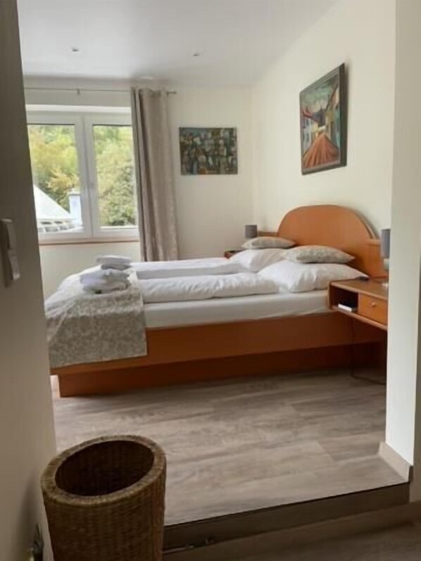 Un dormitorio con cama doble, mesita de noche, lámpara, un cuadro en la pared y ventana con cortinas.