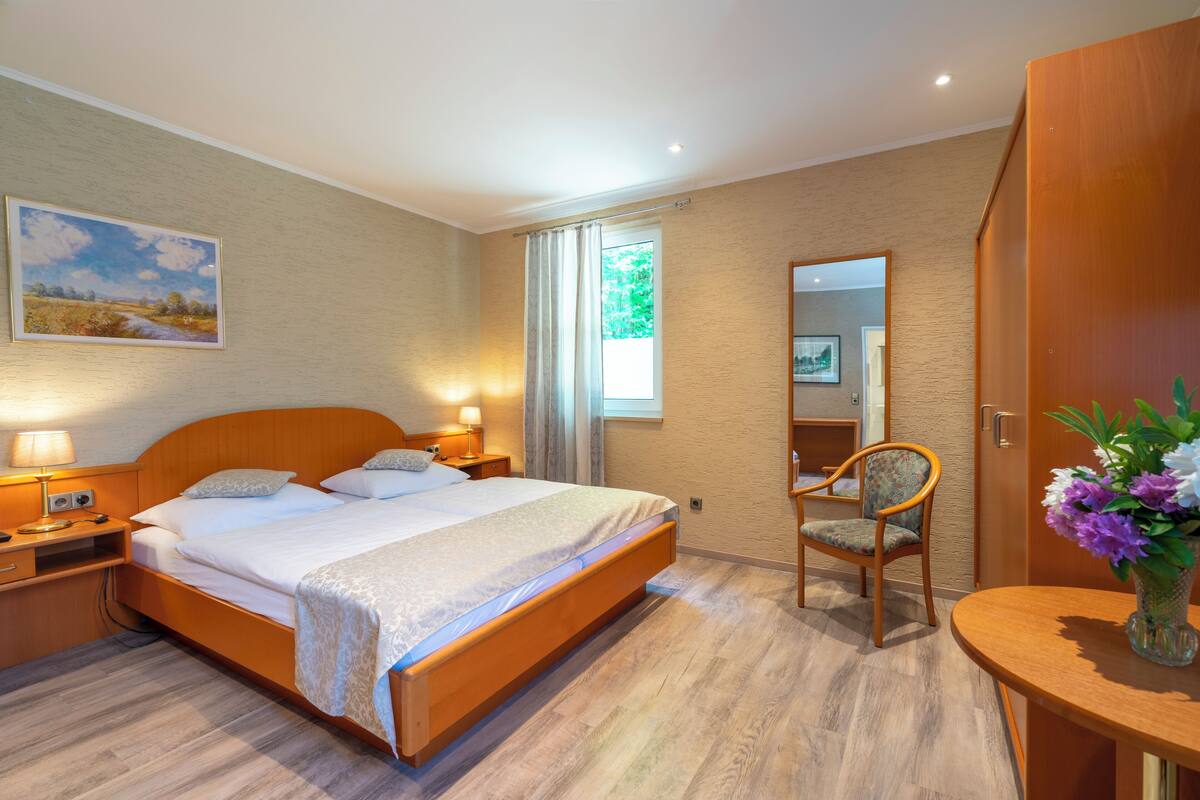Una habitación de hotel con una cama grande, lámparas de noche, una silla, una mesita con un florero y una ventana con cortinas.