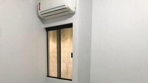 A small, single-bed room with a window, an air conditioning unit above it, and a small table with a book. - Hotel Oliveira 44 (Goiânia)