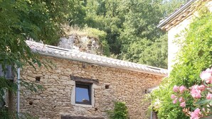Um edifício rústico de pedra com uma pequena janela e porta aberta, rodeado por vegetação e um caminho de pedra.