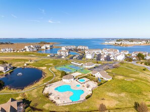 Ett flygbildsbild av ett bostadsområde med en stor swimmingpool, flera flerfamiljshus och en vattenyta i bakgrunden.