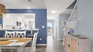 A modern kitchen with white cabinets, a blue accent wall, and a dining area with white chairs and a wooden table. - Bethany Bay Hideaway: Spacious, Modern 4 bed, 4 bath condo, water & wetland view (Ocean View)