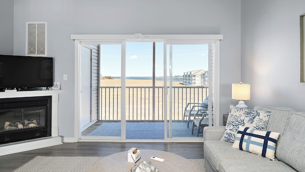 A living room with a fireplace, a television, a sofa with patterned cushions, and a view of the beach through large sliding doors. - Bethany Bay Hideaway: Spacious, Modern 4 bed, 4 bath condo, water & wetland view (Ocean View)