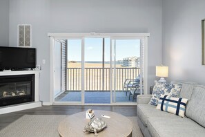 A living room with a fireplace, a television, a sofa with patterned cushions, and a view of the beach through large sliding doors. - Bethany Bay Hideaway: Spacious, Modern 4 bed, 4 bath condo, water & wetland view (Ocean View)