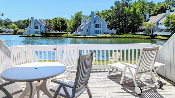A deck with white chairs and a table overlooking a body of water with houses in the background. - Ground-Floor Sea Colony Condo. Pond Views. Resort Amenities! (Bethany Beach)